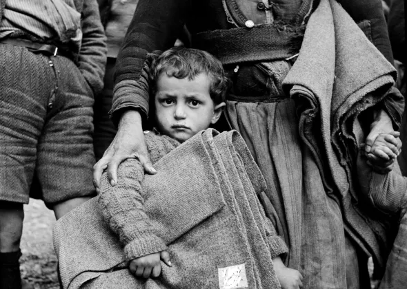 Circa 1950 in the north-western town of Castoria, a boy holds a folded UNICEF-supplied blanket, clutching the skirt of a woman who has one hand on his shoulder and the other holding the hand of another child, with another blanket under her arm.