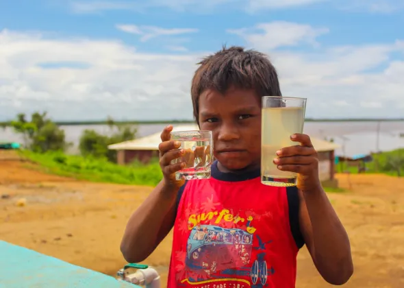 A child looking at the camera during the installation of a tank of 45,000 liters, which will provide access to potable water to an estimated 1,500 people per month, among which 500 indigenous people of the Warao ethnic group stand out, Cambalache Community, Bolívar state, on July 20, 2020. In his right hand, the child holds water drawn from the newly installed water tank; in his left, he holds water from the river, which had been the community's water source prior to the tank installation.